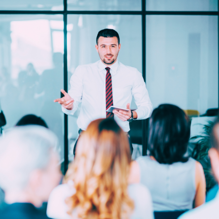 Treinamento: homem usa camisa branca e gravata. Está de pé fazendo uma palestra para o público que está sentado a sua frente.