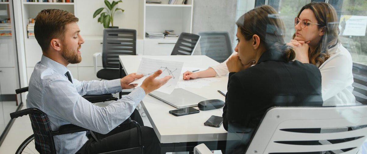Inclusão: 2 mulheres e 1 homem em cadeira de rodas conversam em uma sala de reunião.