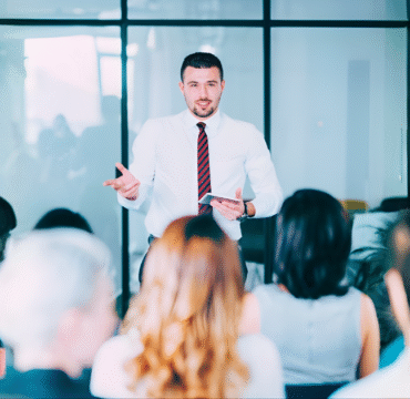 Treinamentos: homem usa camisa branca e gravata. Está de pé fazendo uma palestra para o público que está sentado a sua frente.
